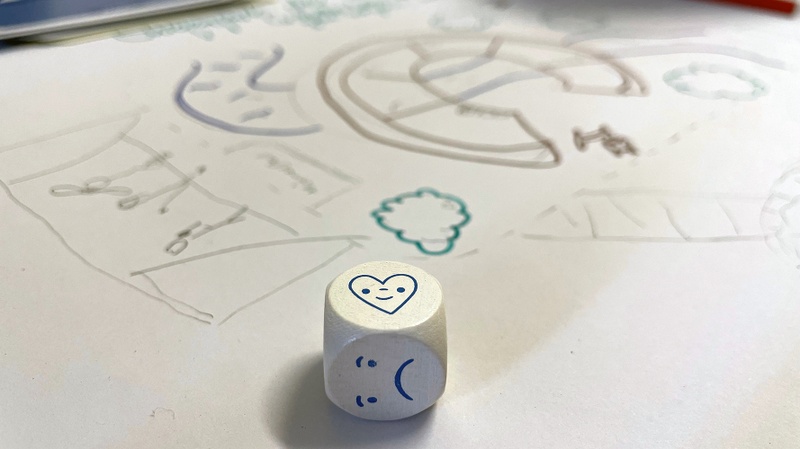 Ein Würfel mit einem lachenden Herz und einem traurigen Smiley liegt auf einer Zeichnung