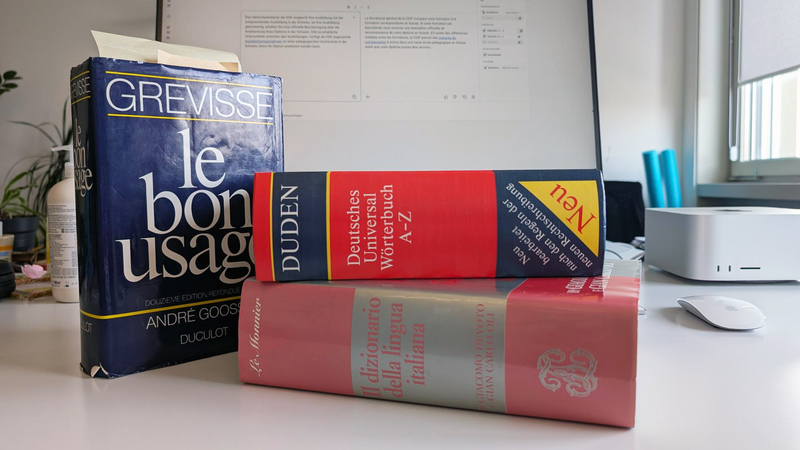 Trois gros dictionnaires (français, allemand et italien) sont posés sur un pupitre ; à l'arrière-plan, on aperçoit un écran sur lequel est ouvert DeepL